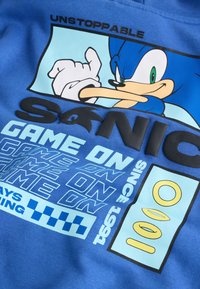 Postava Sonica na modré látce s textem "UNSTOPPABLE," "SONIC GAME ON," "SINCE 1991," a grafikou zlatých prstenů.