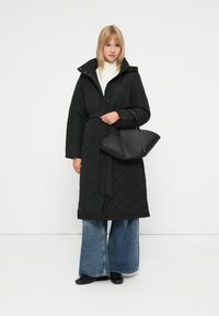 Juodas siuvinėtas ilgas paltas su diržu liemeniui, turintis gobtuvą. Suderinta su plačiomis džinsinėmis kelnėmis ir struktūrizuotu juodu rankiniu krepšiu.