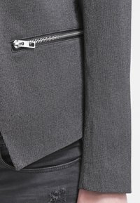 Giacca grigia con texture e tasca con zip. Presenta bordi sagomati e una leggera lucentezza. Il materiale sembra tessuto, abbinato a pantaloni neri consumati.