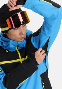 Giacca da snowboard blu e nera con accenti gialli, ventilazioni con zip e polsini regolabili. Il modello indossa occhiali da sci riflettenti e un berretto nero.