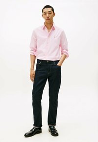 Lichtroze overhemd met knopen, donkere spijkerbroek en zwarte instappers. Het overhemd heeft opgerolde mouwen; de jeans hebben een klassieke pasvorm met subtiele steken.