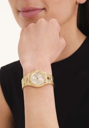 Muñeca con un reloj de eslabones de metal dorado, con esfera redonda y números romanos, levantada cerca de la barbilla de una mujer con blusa negra.