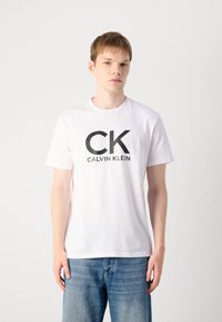 Witte katoenen t-shirt met een groot zwart "CK" logo en de tekst "CALVIN KLEIN". Korte mouwen en een ronde hals, gecombineerd met een blauwe spijkerbroek.