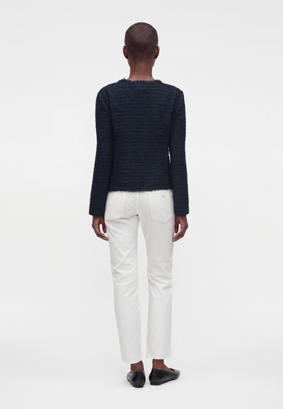 Personne de dos, portant un haut à manches longues texturé bleu marine, un pantalon blanc et des chaussures plates noires sur un fond blanc uni.