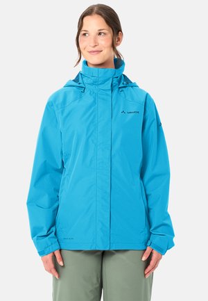 Frau trägt eine leuchtend blaue, wasserdichte Vaude-Jacke mit hohem Kragen und verstellbaren Ärmelbündchen, steht vor einem schlichten Hintergrund.