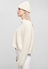 Vit stickad kort cardigan med ett texturerat mönster, parat med en matchande vit mössa. Outfiten har en avslappnad passform och sänkt axelsöm.
