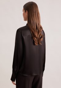 Donna con lunghi capelli castani mossi, che indossa una blusa nera di satin a maniche lunghe e pantaloni scuri, di spalle su uno sfondo neutro.