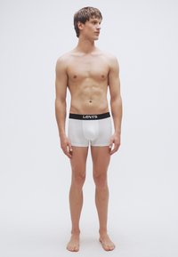 Valkoiset bokserialushousut, joissa on musta Levi'sin vyötärönauha, päällään seisova miesmalli ilman paitaa, yksinkertaisella taustalla.