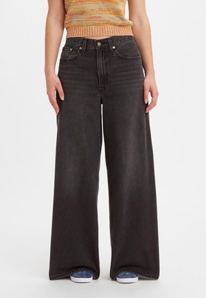 Personne portant un jean noir taille haute à jambes larges, un top court en maille multicolore et des baskets bleues sur un fond blanc uni.