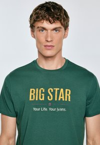 Roheline puuvillane t-särk, millel on suur kollane tekst "BIG STAR" ja väiksem valge tekst "Teie Elu. Teie Jeans." Ümmargune kaelus ja lühikesed varrukad.