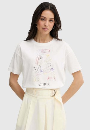 Donna con capelli scuri che indossa una T-shirt bianca con disegni colorati a forma di lettera A e gonna beige a vita alta con cintura.
