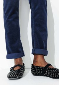 Ballerines en suede noir avec des œillets métalliques. Un pantalon en velours côtelé bleu navy retroussé révèle les chaussures, mettant en valeur leur forme arrondie et leur bride.