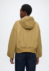 Mustársárga kapucnis dzseki rugalmas szegéllyel, laza szabással és patentos részletekkel az ujjakon. Sima textúrájú; minimalista dizájn, hátulnézet.