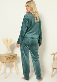 Femme en haut à manches longues en velours vert et pantalon de détente, se tenant pieds nus à côté d'une table en bois avec des fleurs séchées beiges.