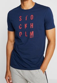 Mand iført navyblå t-shirt med lodret rød tekst, der lyder "Stockholm", og grå joggingbukser med striber i siden, hånd i lommen.