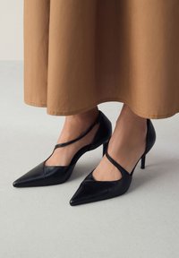 Czarne szpilki z ostrym noskiem i wycięciami. Wykonane z gładkiej skóry, z cienkim obcasem i paskiem skrzyżowanym na stopie.