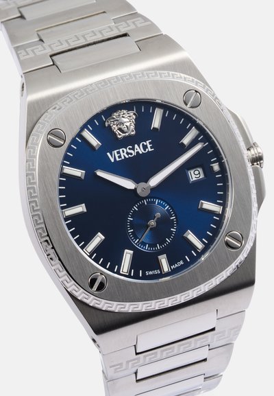 Reloj de acero inoxidable con esfera azul, marcadores de hora plateados, pantalla de fecha y un logo de Medusa. Presenta un bisel texturizado y una pulsera con patrón.