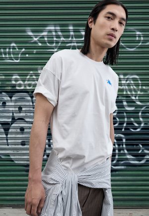 White cotton t-shirt with rolled sleeves, featuring a small blue logo on the chest. Striped fabric tied around the waist, paired with brown pants.