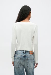Persona con cabello largo y rizado, vistiendo un suéter blanco y jeans azules descoloridos, de pie mirando hacia atrás contra un fondo claro y liso.