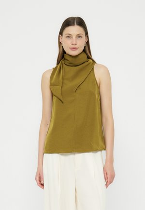 Femme portant un top sans manches vert olive avec un détail d'écharpe col montant et un pantalon blanc à jambes larges, debout devant un fond uni.