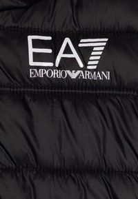 Černá vycpaná bunda se prošívaným designem. Na látce výrazné bílé loga EA7 a Emporio Armani. Hladká, lesklá textura.
