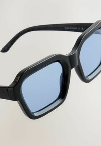 Ochelari de soare negri cu o ramă octagonală, având lentile colorate în nuanțe de albastru și un finisaj lucios. Brațele sunt elegante și arcuite pentru un aspect modern.