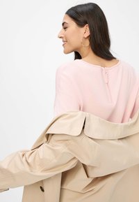 Lichtroze sweater met een ritsdetail op de rug, gedragen met een tan trenchcoat over de schouder. Soepele textuur en casual ontwerp.