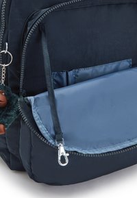 Marinblå ryggsäck med en texturerad tyg, utrustad med en blå dragkedjeficka, silverfärgad metall och en pälsig charm fäst vid dragkedjan.