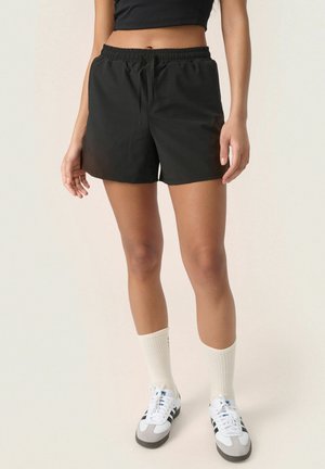 Frau trägt schwarze Shorts mit Kordelzug, weiße Sportsocken, schwarz-weiße Turnschuhe und ein schwarzes kurzes Oberteil, steht vor hellem Hintergrund.