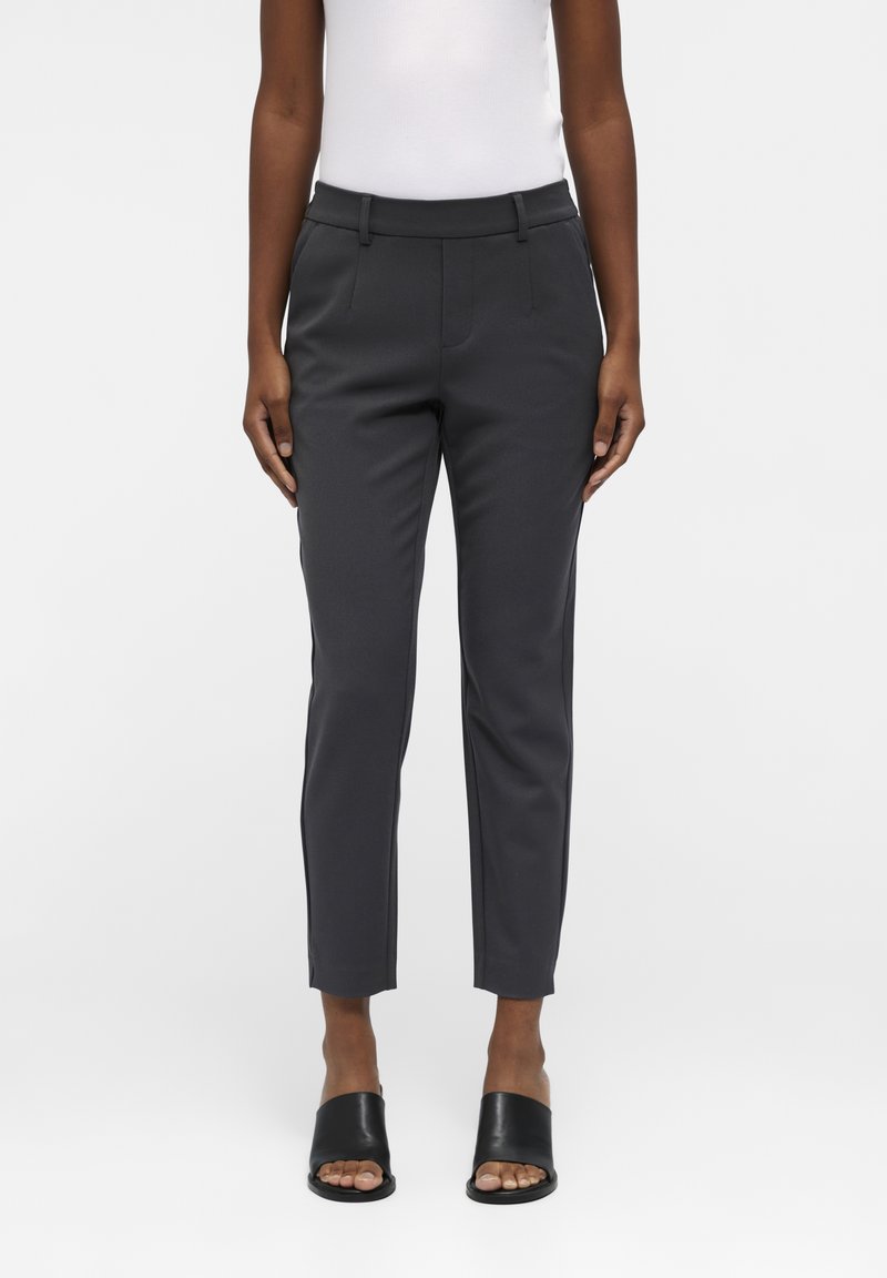 Pantalons taillés gris foncé avec une coupe slim, devant plat et poches latérales. Le matériau semble lisse avec une légère brillance. Associés à des sandales à glissière noires.