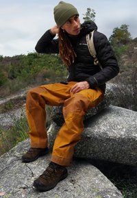 Czarna kurtka puchowa, oliwkowa czapka, pomarańczowe spodnie z brązowymi akcentami i brązowe buty trekkingowe. Siedzi na kamieniu na świeżym powietrzu.