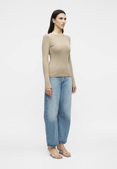 Γυναίκα στέκεται, φορώντας μπεζ ριπ μακρυμάνικη μπλούζα, φαρδιά ανοιχτό μπλε τζιν και μπεζ σαγιονάρες σε ένα απλό λευκό φόντο.