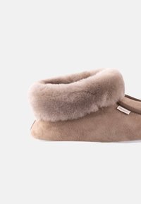 Pantofole in suede tan con un morbido polsino in pelliccia sintetica beige e suola bianca. Presenta un piccolo etichetta sul lato. Design pulito e semplice.