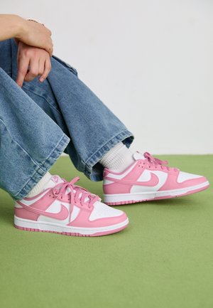 Person siddende med sammenlagte hænder, iført hvide sokker, blå jeans og lyserøde og hvide Nike-sneakers på grøn gulv.