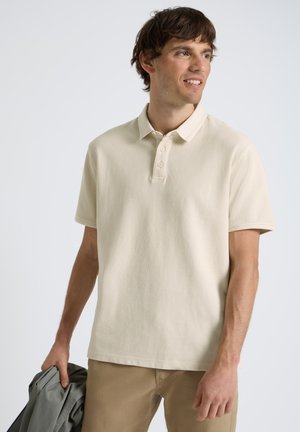 Młody mężczyzna w beżowej koszulce polo i khaki spodniach, trzyma szarą kurtkę, patrzy na bok na neutralnym tle.