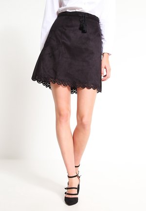 Kobieta ubrana w czarną mini spódniczkę zamszową z falowanym brzegiem i paskiem z frędzlami, w połączeniu z czarnymi szpilkami na kostkę i białą bluzką.