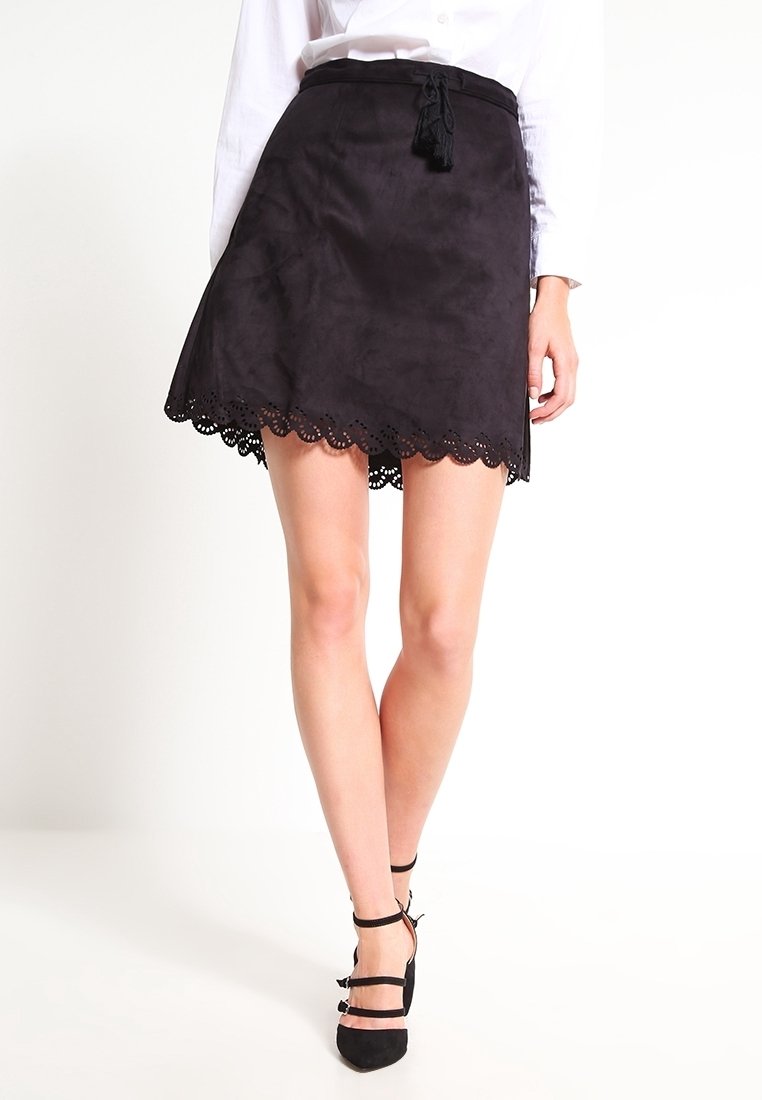 Femme portant une mini-jupe en daim noir à ourlet festonné et ceinture à franges, associée à des talons noirs à brides à la cheville et un chemisier blanc.