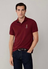 Bordová polokošeľa s golierom, s malým vyšívaným vzorom na ľavej strane hrudníka. Krátke rukávy a hladká textúra, kombinované s tmavými džínsami.