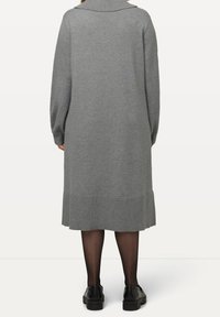 Femme portant une longue robe en tricot grise avec un large col, des collants noirs transparents et des mocassins noirs, vue de dos.