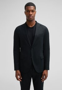 Schwarzer Blazer aus strukturiertem Stoff, mit Ein-Knopf-Verschluss, Reverskragen, Fronttaschen, kombiniert mit einem dunklen Pullover darunter.