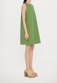 Grünes ärmelloses Kleid mit rundem Ausschnitt und A-Linien-Silhouette. Kombiniert mit beigen Plateausandalen mit Holzabsatz und offenem Zeh.