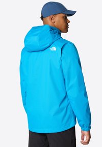 Modrá nepromokavá bunda s kapucí, elastickými manžetami a logem na zadní straně. Hladký materiál, lehká tkanina, navržena pro venkovní použití.