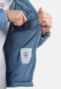 Giacca isolante blue con una texture liscia; dotata di chiusura con zip, tasca interna e una patch con logo sul lato sinistro.