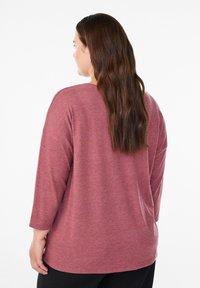 Woman with long brown hair wearing a loose, long-sleeve dusty rose top and black pants, shown from behind against a white background.