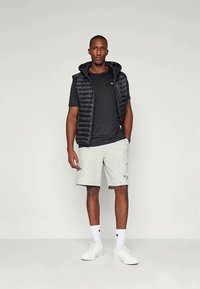 Juodas pūkinis liemenė virš juodo marškinėlio, derinama su šviesiai pilkomis šortais. Aksesuarai apima baltas kojines ir sportbačius, minimaliai matomas ženklas.