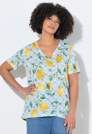 Kvinde med krøllet hår iført en V-hals trøje med gul citron- og grønt løvmønster, kombineret med en blå nederdel, stående mod en hvid væg.