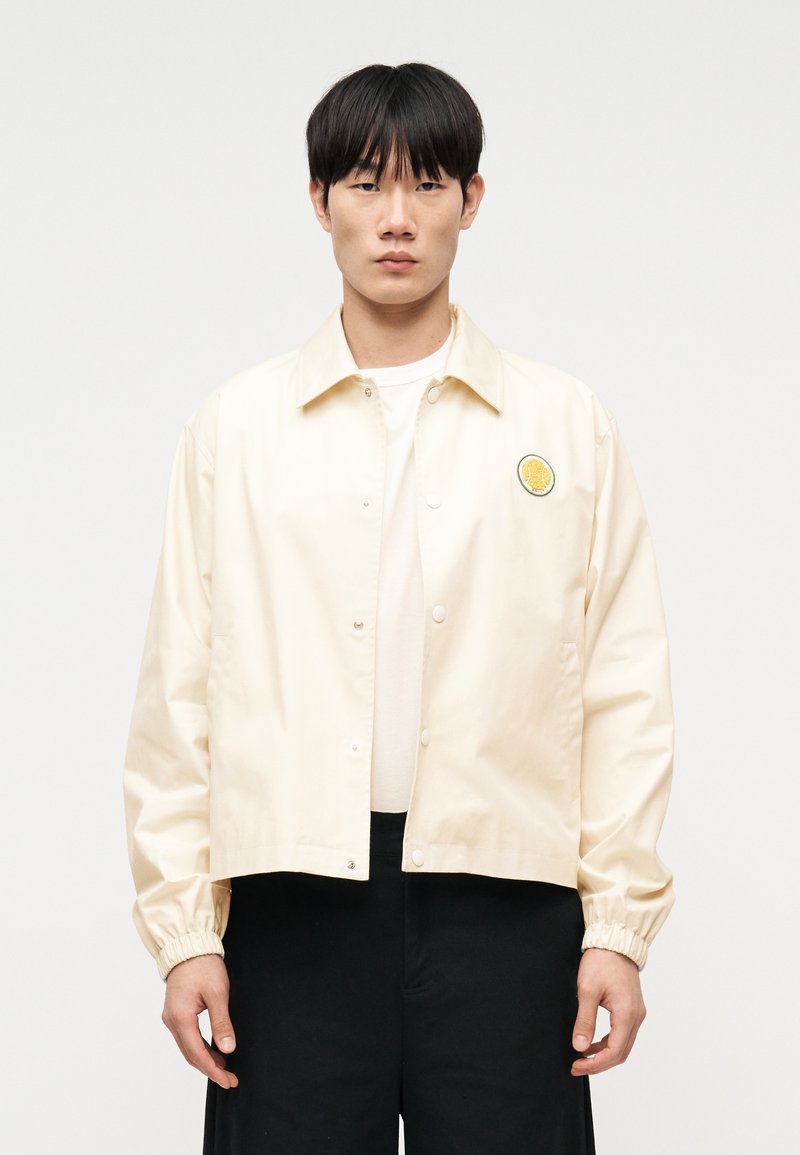 Νεαρός άνδρας που φοράει κρεμ μπου φάν με ελαστικά μανίκια και κίτρινο λογότυπο, σε συνδυασμό με λευκό πουκάμισο και μαύρο παντελόνι, στέκεται μπροστά σε λευκό φόντο.