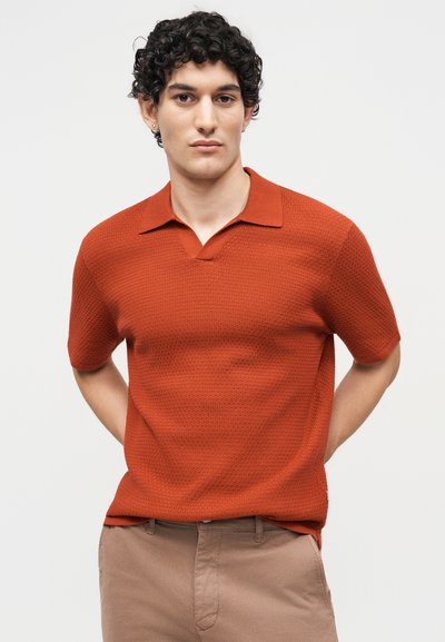 Jeune homme aux cheveux bouclés portant un polo orange texturé à manches courtes et un pantalon beige, debout, les mains dans le dos.