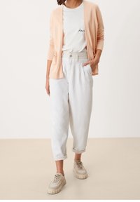 Jasnopomarańczowy sweter rozpinany na biały t-shirt z haftowanym detalem, zestawiony z luźnymi białymi spodniami i beżowymi trampkami o fakturowanych podeszwach.