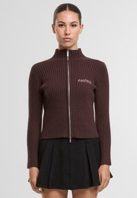 Bordowy prążkowany sweter rozpinany na zamek z wysokim kołnierzem i haftowanym napisem "ANOTHER" na piersi. Noszony z czarną plisowaną spódnicą.